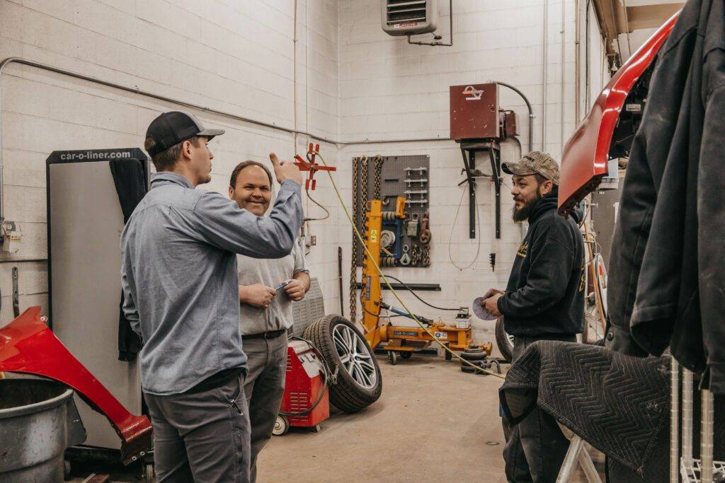 Three men stand in a garage beside a red car