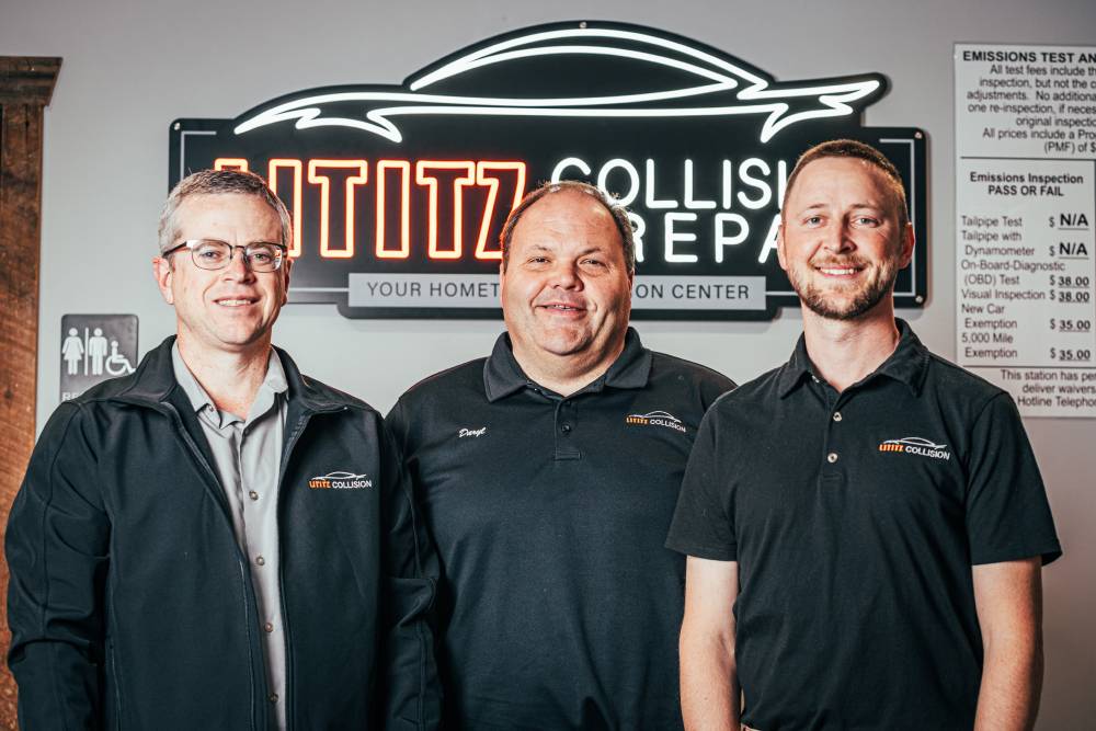 Three certified technicians from Lititz Collision Repair standing in front of a neon sign, showcasing expertise and commitment to quality auto body services.