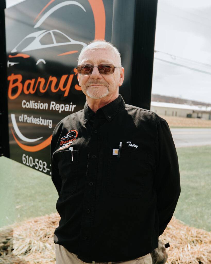 Tony from Barry's Collision Repair, wearing a black shirt with his name tag, standing in front of the business sign.