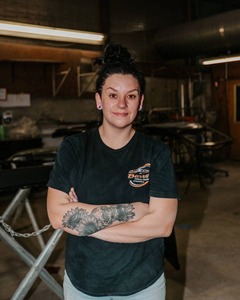Michelle from Barry's Collision Repair, standing confidently with crossed arms in the paint department, showcasing her tattooed arm and wearing a branded shirt.